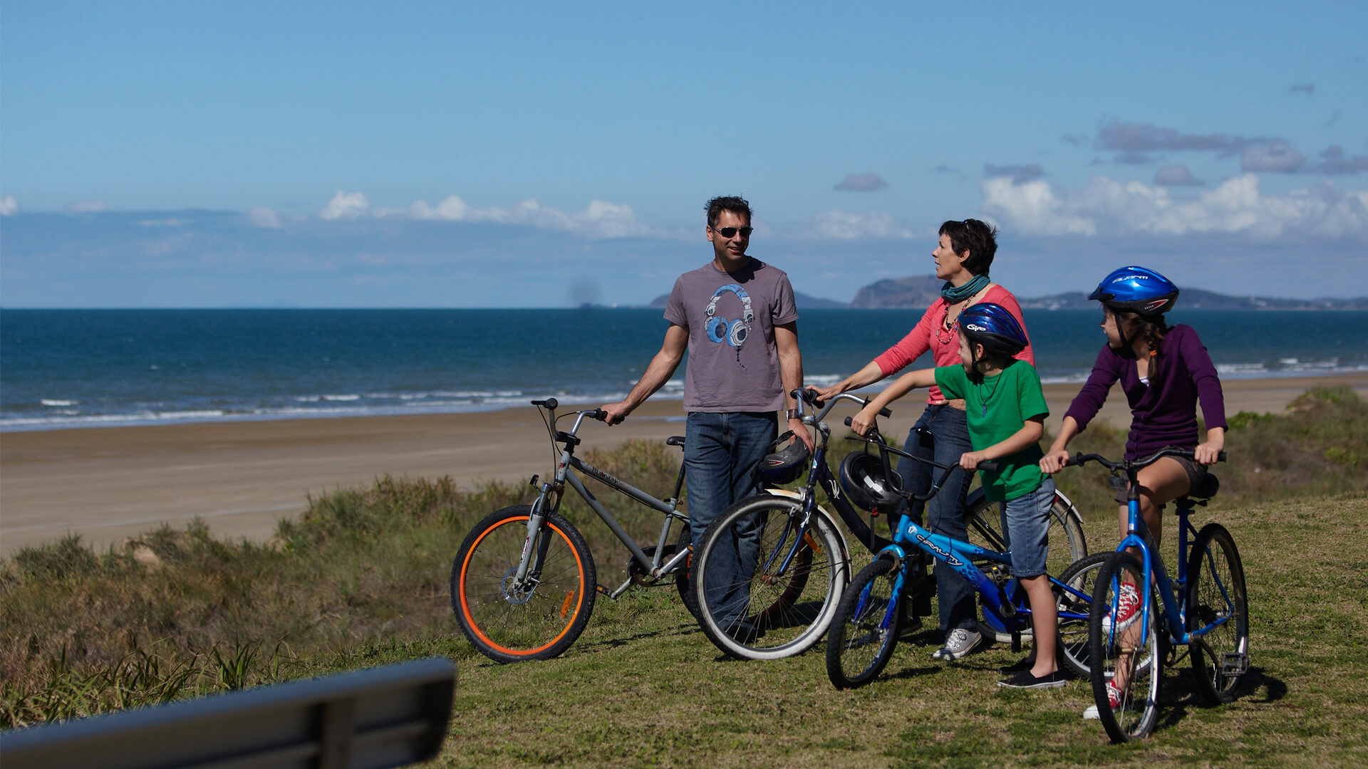 Image of two adults and two children with bicycles standing on grass in front of the ocean. 